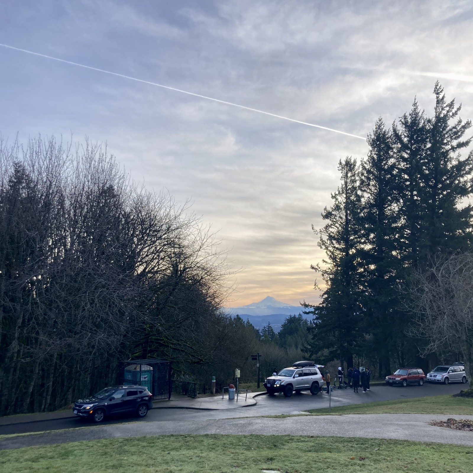 View from Council Crest Park toward Mt. Hood, which is visible in a mostly-clear sky about an hour after sunrise. Mid-altittude clouds are moving in from the south/southwest but these are higher than the mountain. The clouds glow warmly with light reflected from the sunrise, but the scene is chilly and damp otherwise. A single jet contrail crosses the entire width of the photo. In the foreground about 100 feet distant stand a group of about six people next to an open SUV. They are either setting up or tearing down a small but complicated photography rig, tripods and reflectors and such. They have two electric scooters with them. I would guess they were filming or photographing creative for a marketing campaign, something that happens with relative frequency in this park.