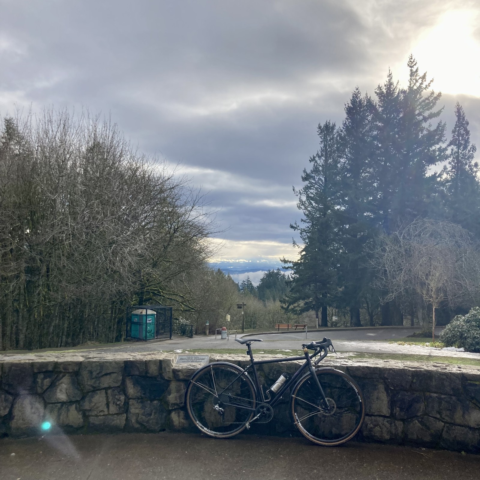View from Council Crest Park toward Mt Hood, the base of which can be discerned behind several layers of clouds, mist, and precipitation. The sky today is…complicated. There is a high deck of broken stratus clouds, through which the sun breaks through in streaks. This layer of clouds obscures the top of Mt. Hood, and a sheet of precipitation falls over the mountain itself. Across the city — so below 500' elevation — an inversion traps pockets of mist. Directly between us and the mountain, clinging mostly to Mt. Hood’s foothills, are a few scattered silvery clouds of precipitation. Many of these clouds glow brightly in the patches of sunlight that break through the top deck. In the foreground, a bicycle leans against  decorative stone wall. A few small pockets of filthy, slushy ice cling to a few spots on the walkways, the remnants of snow from almost two weeks ago. About 100' distant, to the right/south and slightly downhill, is a stand of tall Douglas firs. At approximately the same d