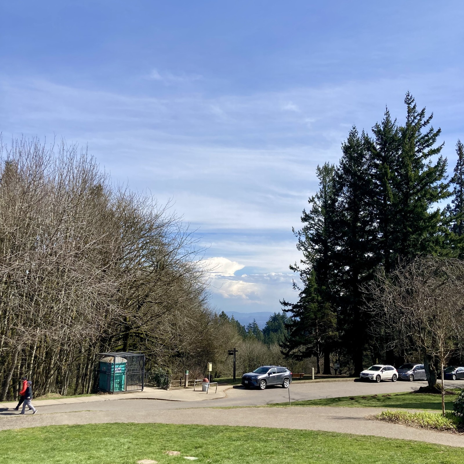 View from Council Crest Park toward Mt. Hood, which huddles under a tall pile of storm clouds. Wisps of high altitude clouds cover the rest of the sky overhead. Clear, sunny, warm! About 50' away and somewhat downslope two hikers walk along the road past a portapotty.
