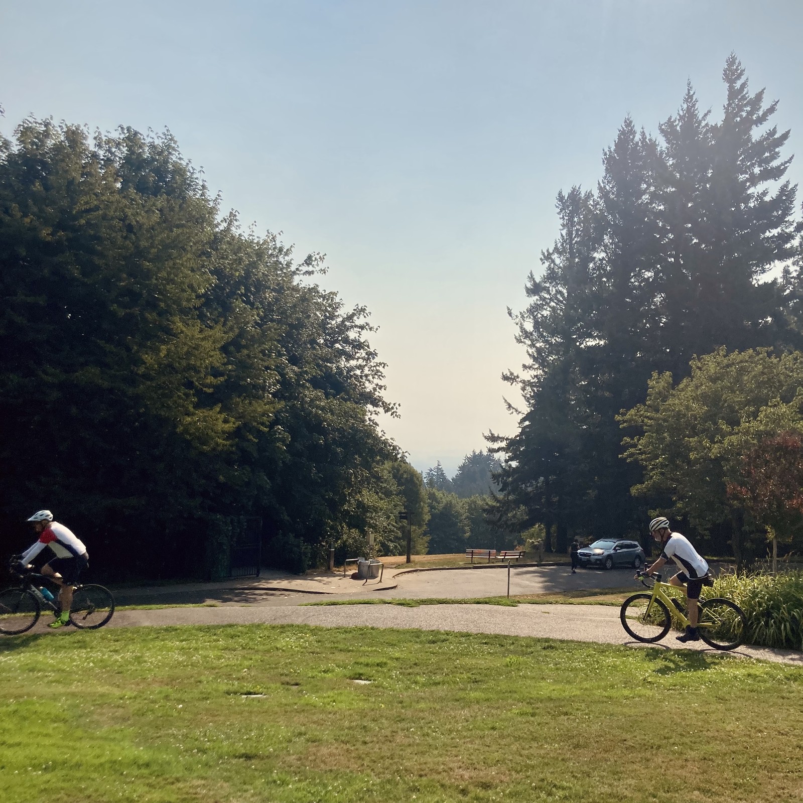Looking toward Mt. Hood from Council Crest park in the late morning. The sky is cloudless but humidity and haze from wildfires obscure visibility to perhaps 20miles, so we cannot see the mountain. The air is visibly yellow, but rich with a wonderful aroma of dried leaves, overripe blackberries, pine resin, and yes a hint of wildfire. In the foreground two middle-aged bike riders ride north along a sidewalk