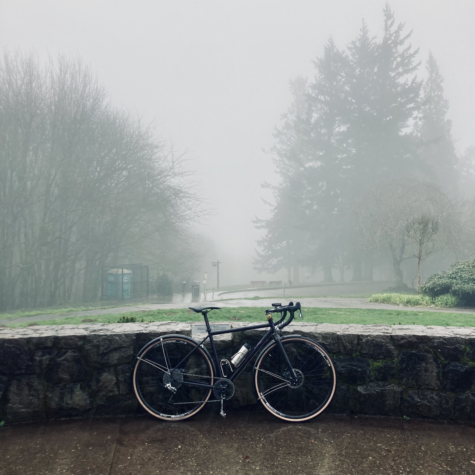A steel bicycle leans against a low stone wall at the top of a hill, in heavy fog and rain