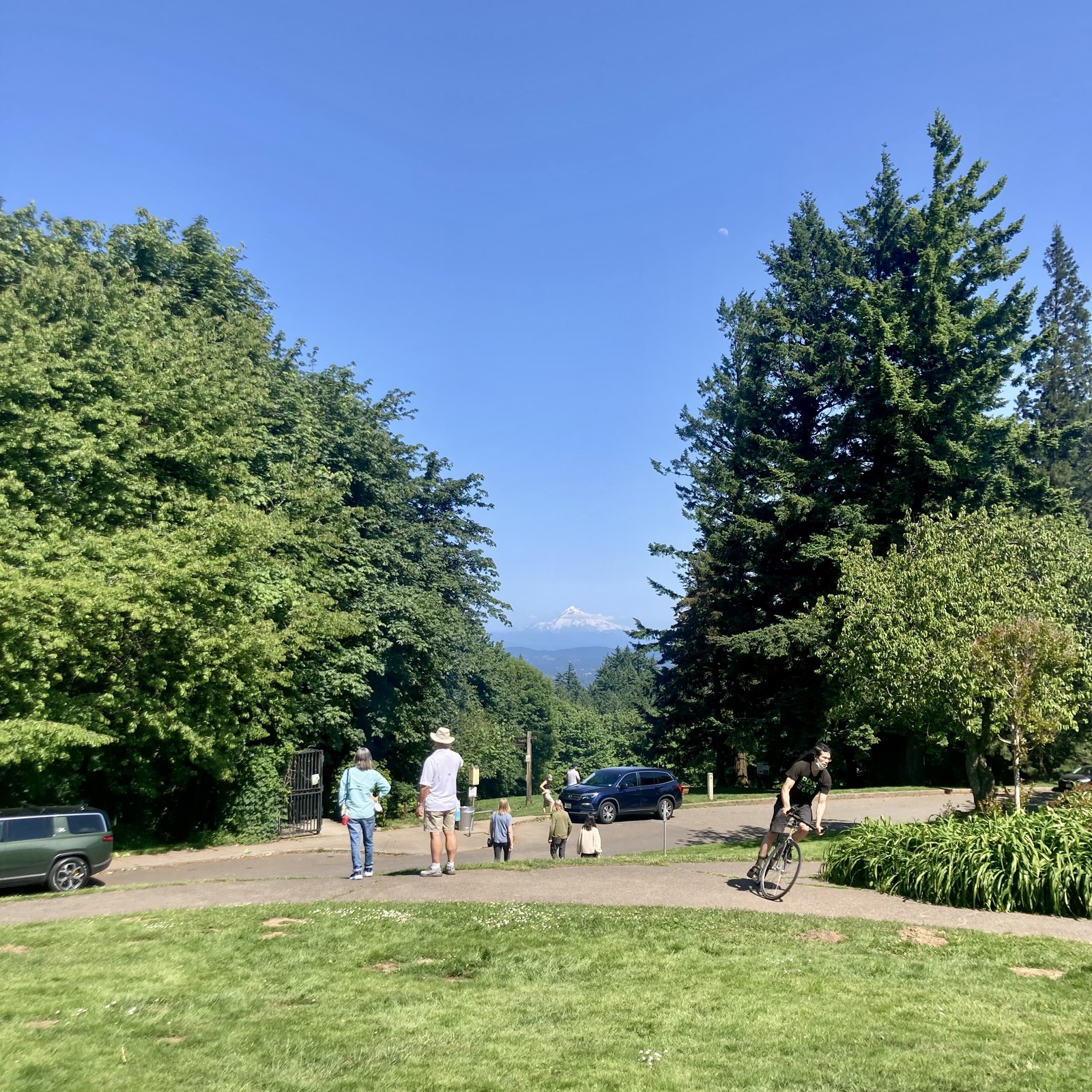 a group of people admire Mt. Hood which is ringed with a layer of clouds under an otherwise spotless sky. A cyclist on a fixed gear bike rides past.