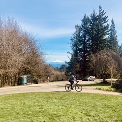 Mt. Hood is snow capped under a shiny bluish grayish sky. About 10m away a man rides a bicycle up the path toward us