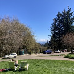 A cheerful sunny Easter Sunday early afternoon at the hilltop park. In the foreground a couple in lawn chairs admire the mountain, which is partly hidden by a few stray clouds. About 50' away another couple lies in the grass, their dog standing over them. A third couple walks along the road about 100' distant. The deciduous trees are just now budding. Bright green grass below and deep blue sky overhead
