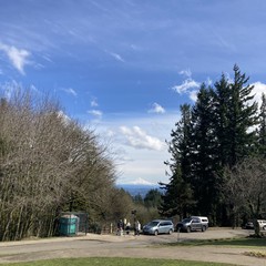 View from Council Crest Park toward Mt. Hood, which is covered with fresh snow and sparkles under a mostly-clear sky. A few high cirrus clouds frame the mountain; a few fluffy clouds are in the mid-sky at a much lower altitude. About 50' away and slightly downhill, a small group of people are looking at the mountain.