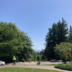 Mt. Hood, brilliant white under a deep blue sky, taken from Council Crest Park. A cyclist in bright yellow scoots his bike up the small hill toward the camera; beyond him a group of three people are looking at the mountain.