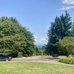 View toward Mt. Hood from Council Crest Park. The sky is hazy with humidity and (at a high altitude) wildfire smoke. The remnants of a marine layer are passing east over the Cascades. Mt. Hood is NOT visible through this layer of clouds. About 100' distant two hikers with small backpacks stand near some park benches