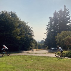 Looking toward Mt. Hood from Council Crest park in the late morning. The sky is cloudless but humidity and haze from wildfires obscure visibility to perhaps 20miles, so we cannot see the mountain. The air is visibly yellow, but rich with a wonderful aroma of dried leaves, overripe blackberries, pine resin, and yes a hint of wildfire. In the foreground two middle-aged bike riders ride north along a sidewalk