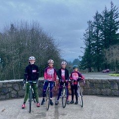Four young women in VALKYR kit pose with their bicycles in front of a low stone wall at the top of the tallest hill in Portland. Behind them are two stands of trees and a gray sky